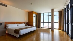 Une chambre d’hôtel dotée d’un grand lit, d’un parquet en bois, d’un bureau intégré et de grandes fenêtres offrant une vue sur la ville.