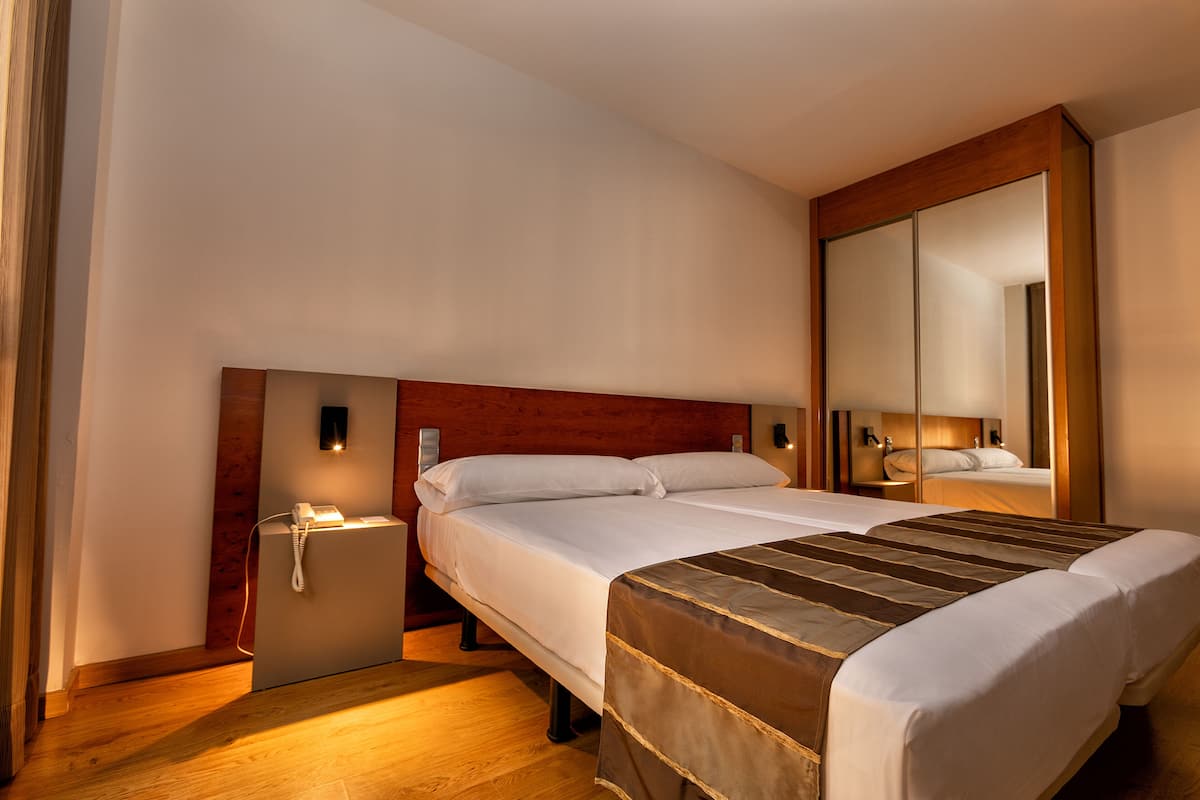 A hotel room with a large bed, a bedside table with a lamp, a mirror, and a wooden floor.