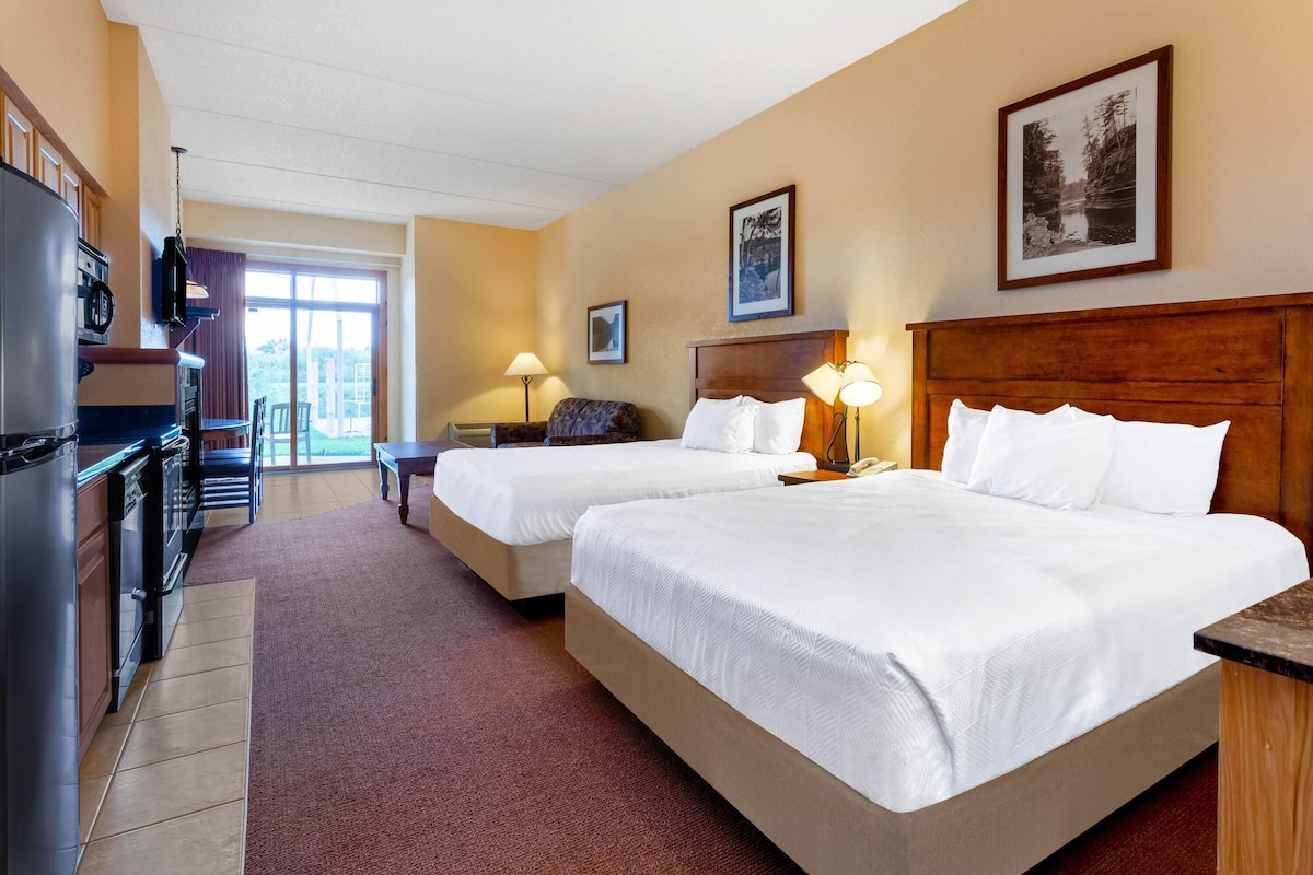 A hotel room with two beds, a desk, a refrigerator, and a dining area with chairs.