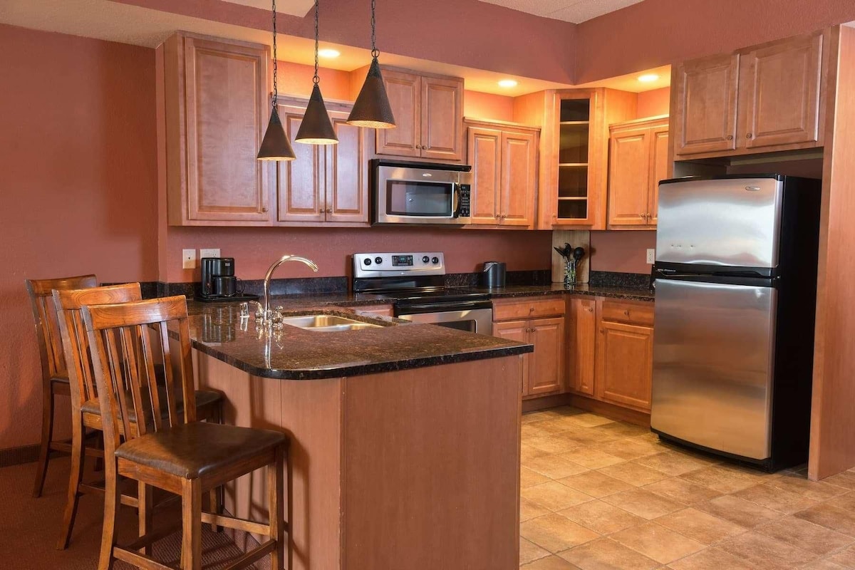 A kitchen with wooden cabinets, a stainless steel refrigerator, a microwave, a stove, and a sink. There are three wooden chairs and pendant lights above the counter.