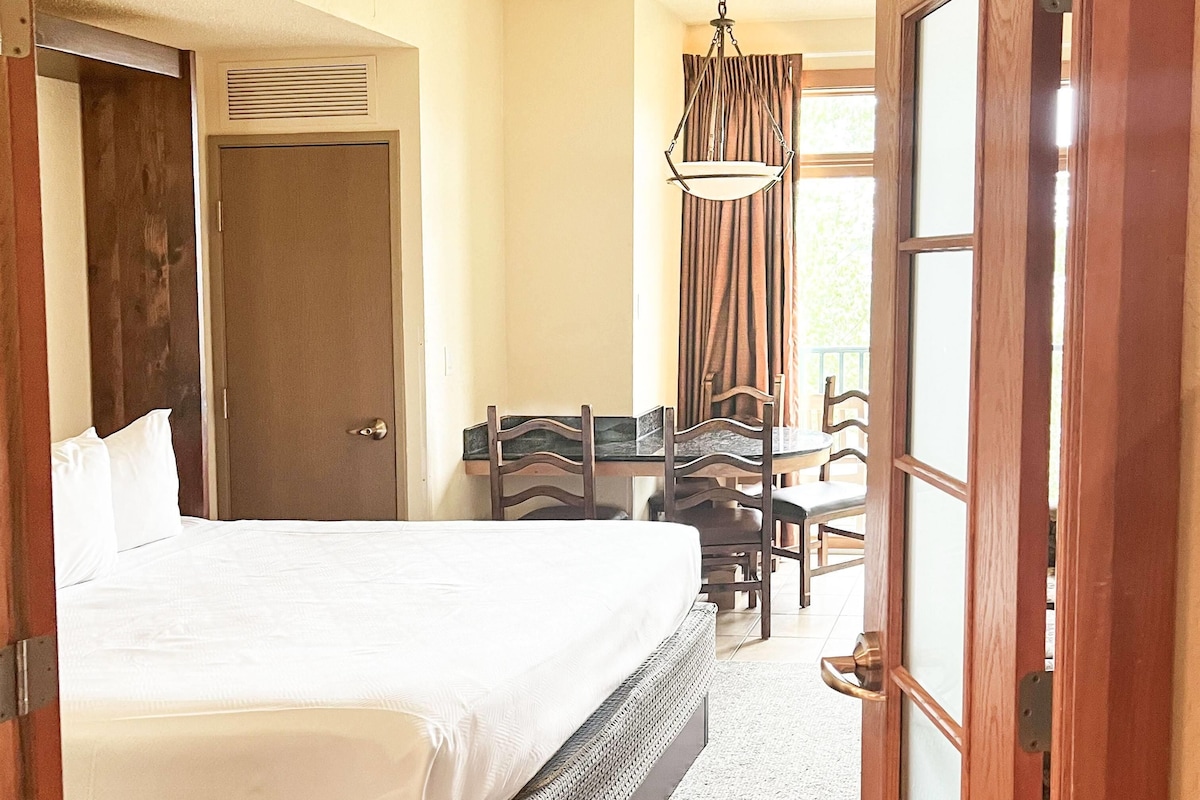 A hotel room with a bed, a desk with chairs, a hanging light fixture, and a door.