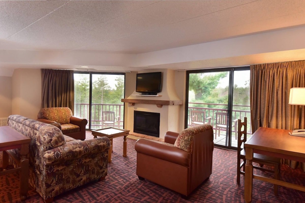 A hotel room with a fireplace, a television, a sofa, a dining table, and a balcony with a view of trees.