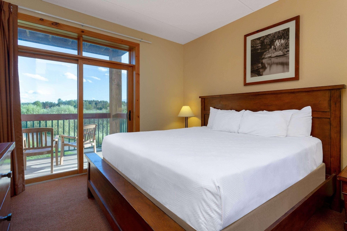 A hotel room with a large bed, a wooden headboard, a nightstand, a lamp, a framed picture on the wall, and a balcony with chairs.