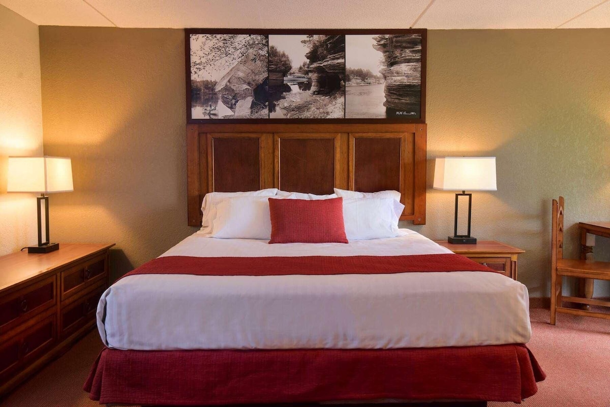 A hotel room with a large bed, two bedside tables with lamps, a wooden headboard, and a framed picture above the bed.