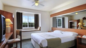 A hotel room with a bed, a desk with a lamp, a chair, a TV, a window with curtains, a ceiling fan, and a mirror. - Microtel Inn & Suites by Wyndham Eagle River/Anchorage Area (Eagle River)