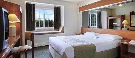 A hotel room with a bed, a desk with a lamp, a chair, a TV, a window with curtains, a ceiling fan, and a mirror.