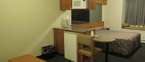 Ein kompaktes Hotelzimmer mit Küchenzeile, einem Einzelbett, einem kleinen Tisch und einem Stuhl.