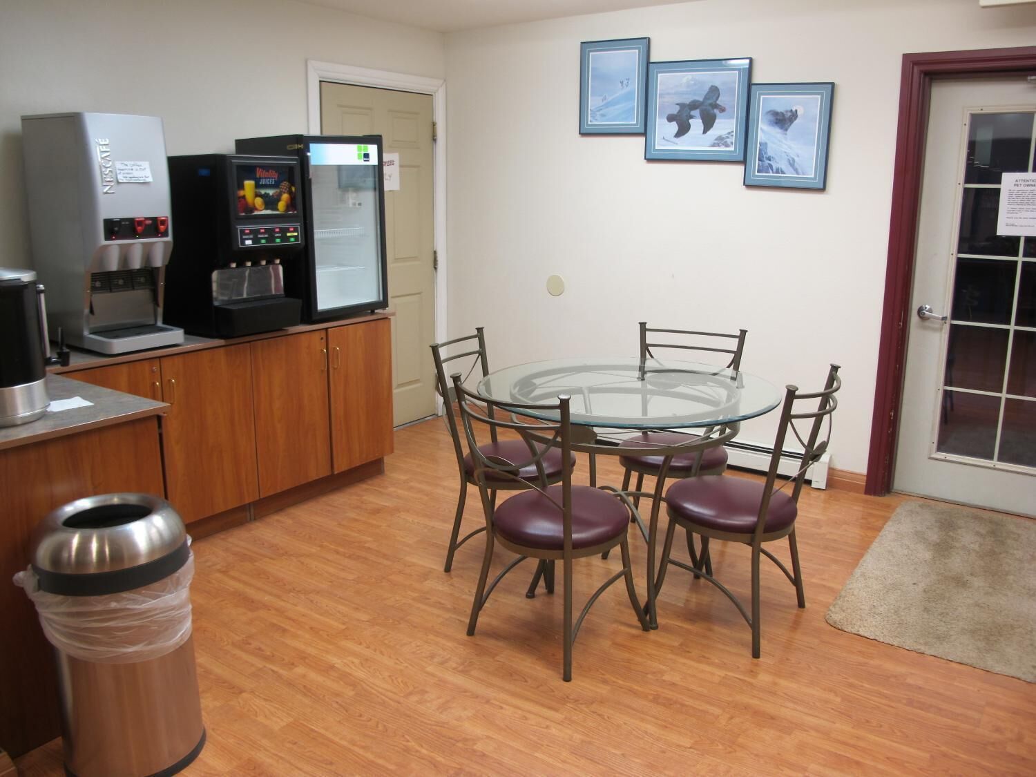 A compact kitchen with a coffee maker, a refrigerator, and a trash bin. There is a glass table with four chairs.