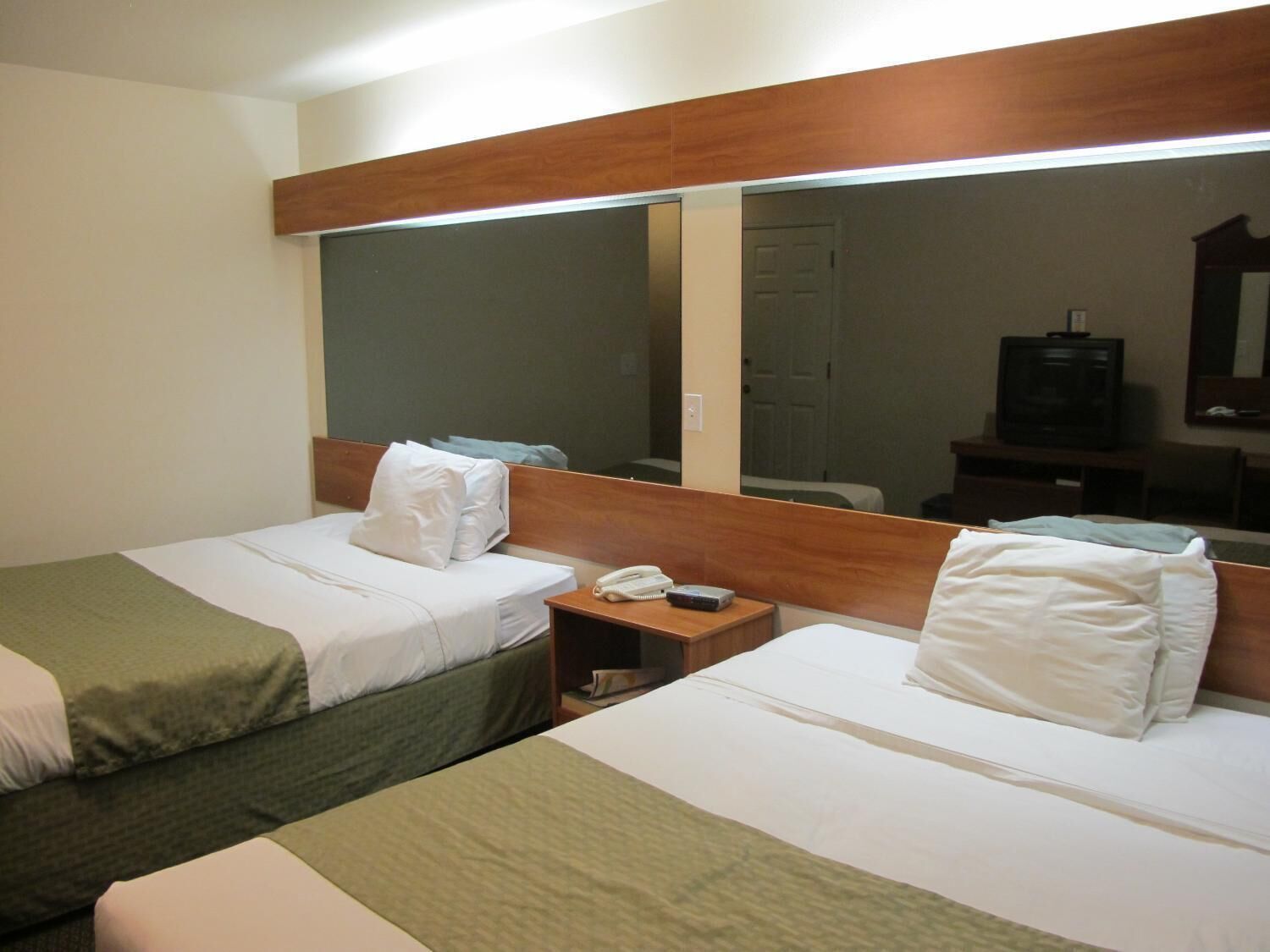 Una camera d'albergo con due letti singoli, un comodino con telefono e telecomando, uno specchio grande e una televisione su un mobile.
