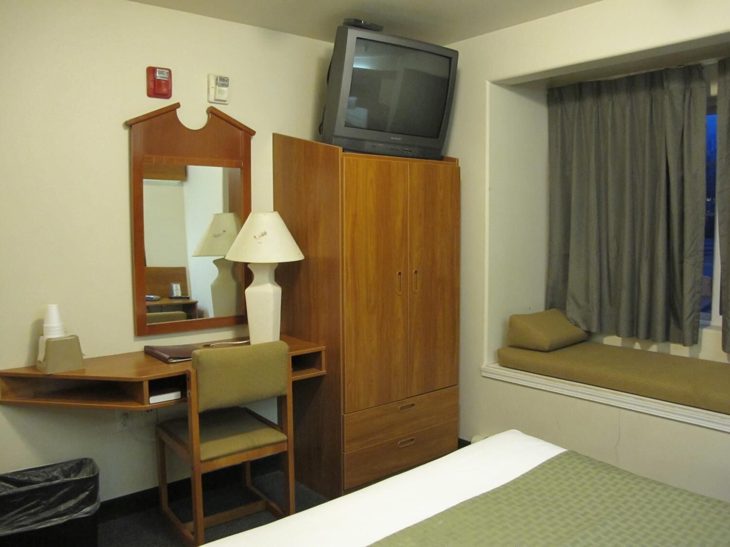 A hotel room with a wooden desk, a chair, a television on top of a wardrobe, a bedside table, and a window with curtains.