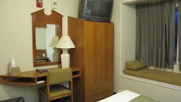 Habitación de hotel con escritorio de madera, una silla, televisor encima de un armario, mesita de noche y ventana con cortinas.