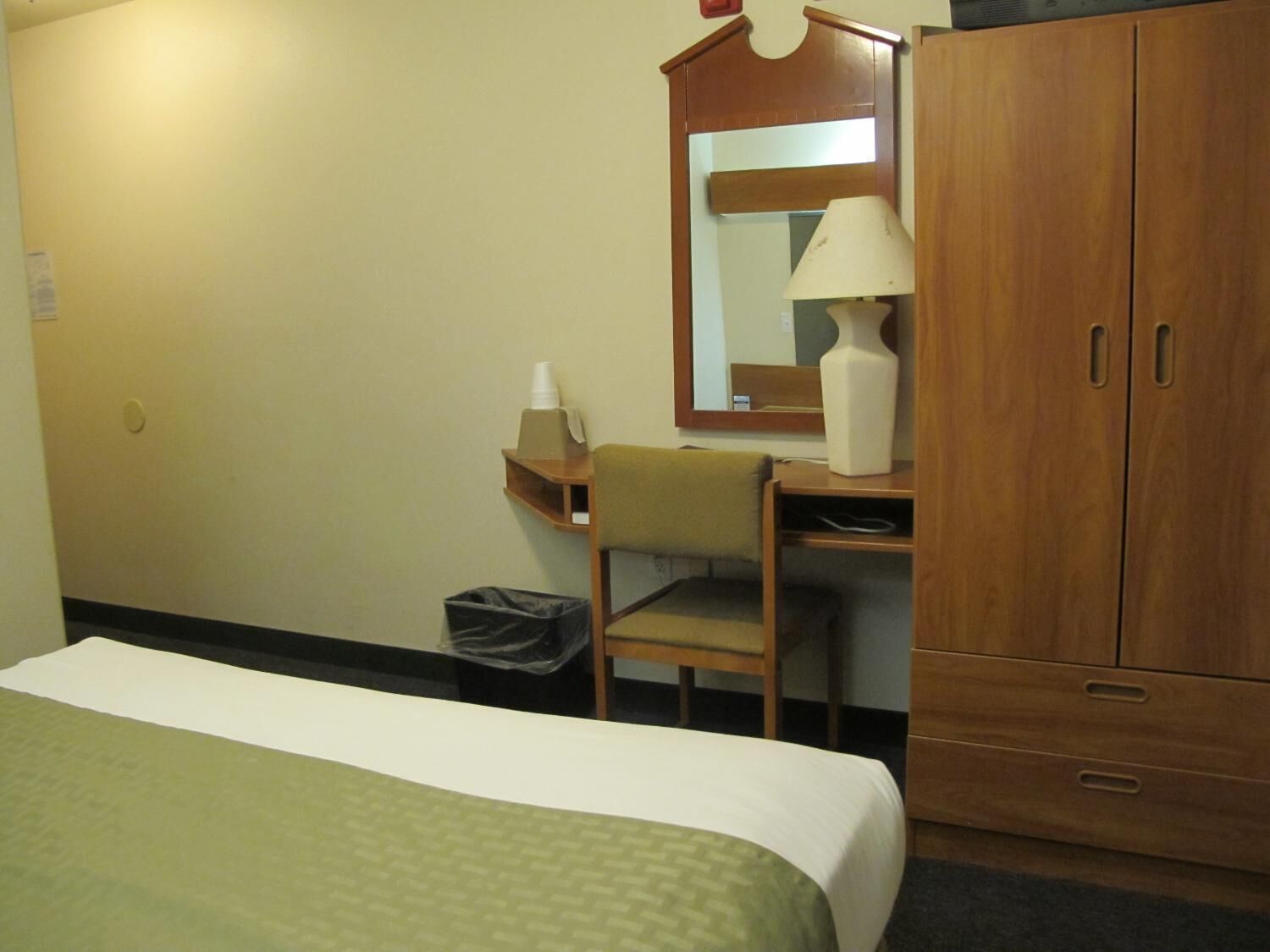 Quarto de hotel com cama, escrivaninha, cadeira, guarda-roupa de madeira, espelho, abajur e lixeira.