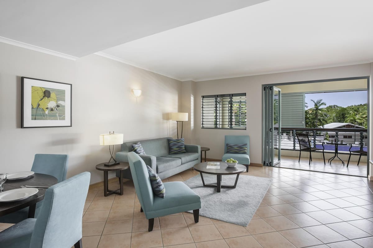 A modern living room with a sofa, armchairs, a coffee table, and a dining area with a table and chairs. There is a balcony with a view of palm trees and a pool.