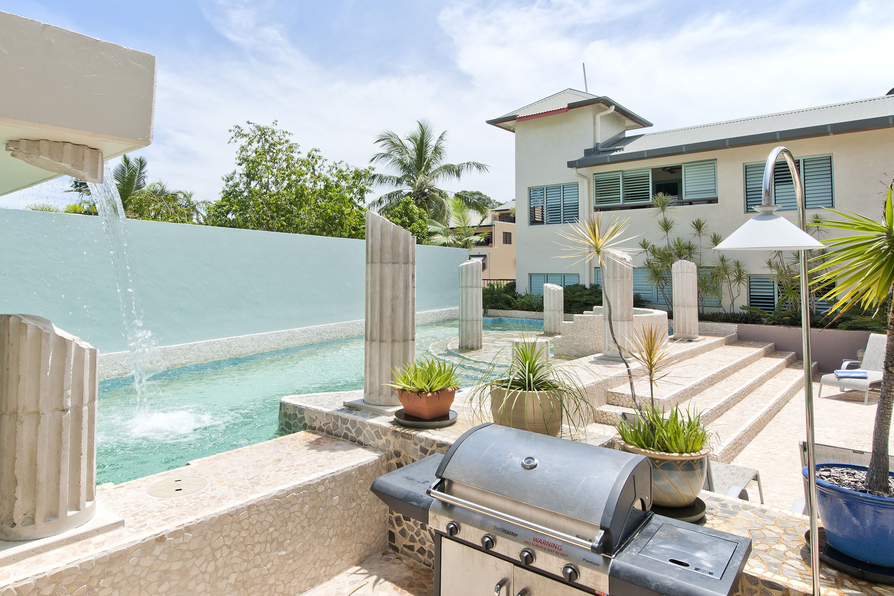 A poolside area with a built-in grill, potted plants, and a view of a modern house in the background.