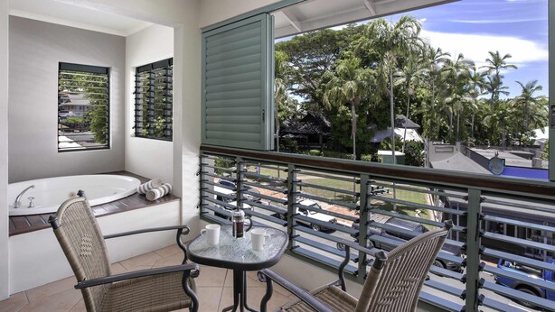 Een balkon met uitzicht op palmbomen en een zwembad, een witte badkuip en rieten stoelen.