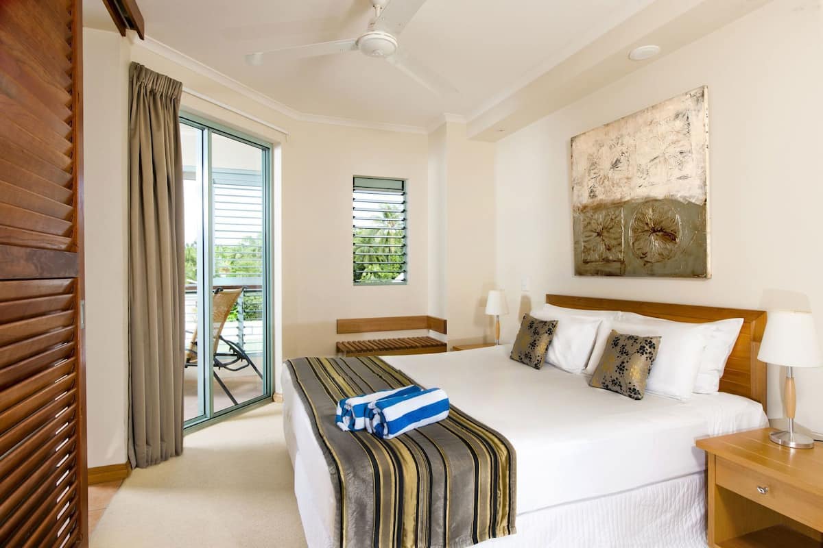A hotel room with a bed, a ceiling fan, a window with blinds, a chair, and a painting on the wall.