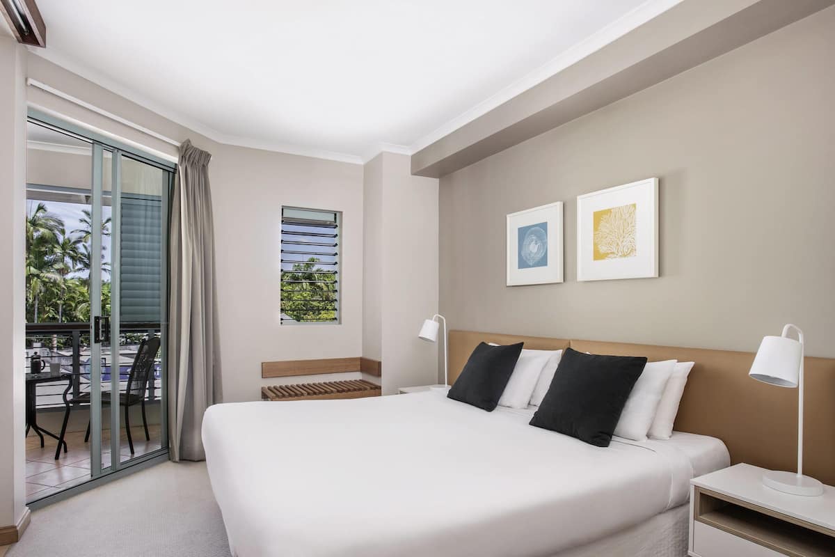 A hotel room with a large bed, a bedside table, a lamp, and a view of the outdoors.