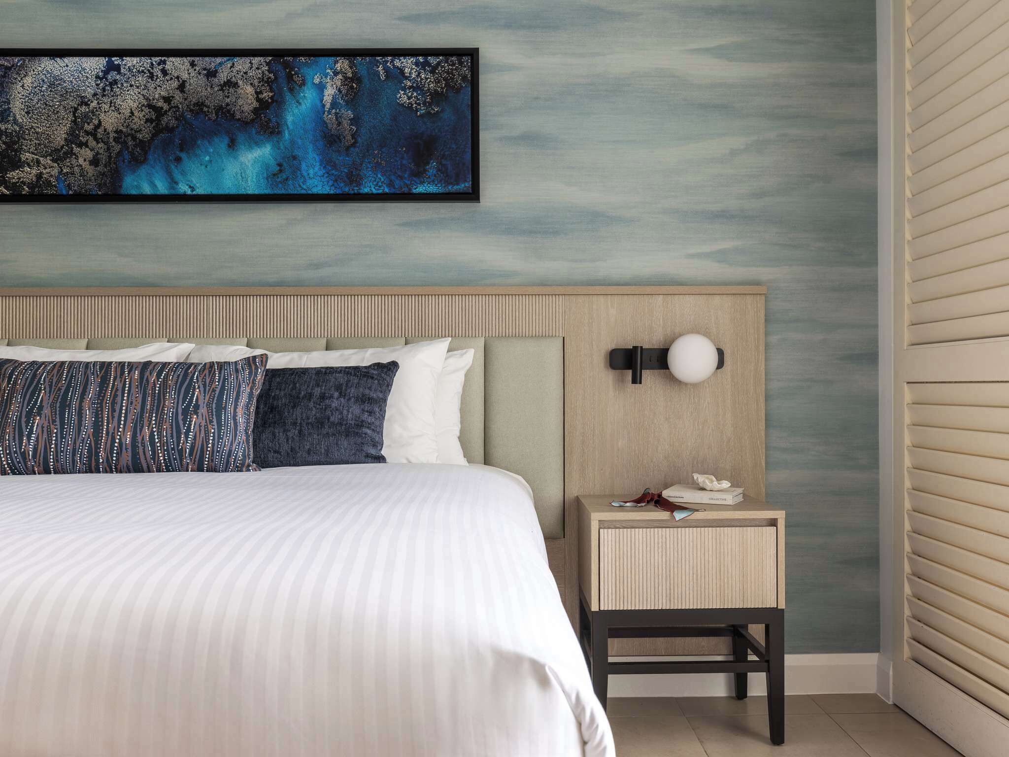 A hotel room with a bed, a nightstand, a wall-mounted lamp, and a large abstract painting.