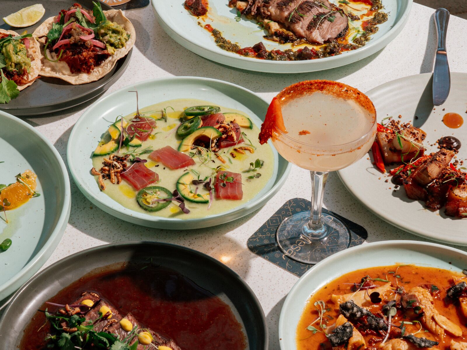 A variety of dishes on a table, including tacos, a soup with avocado, and a cocktail.