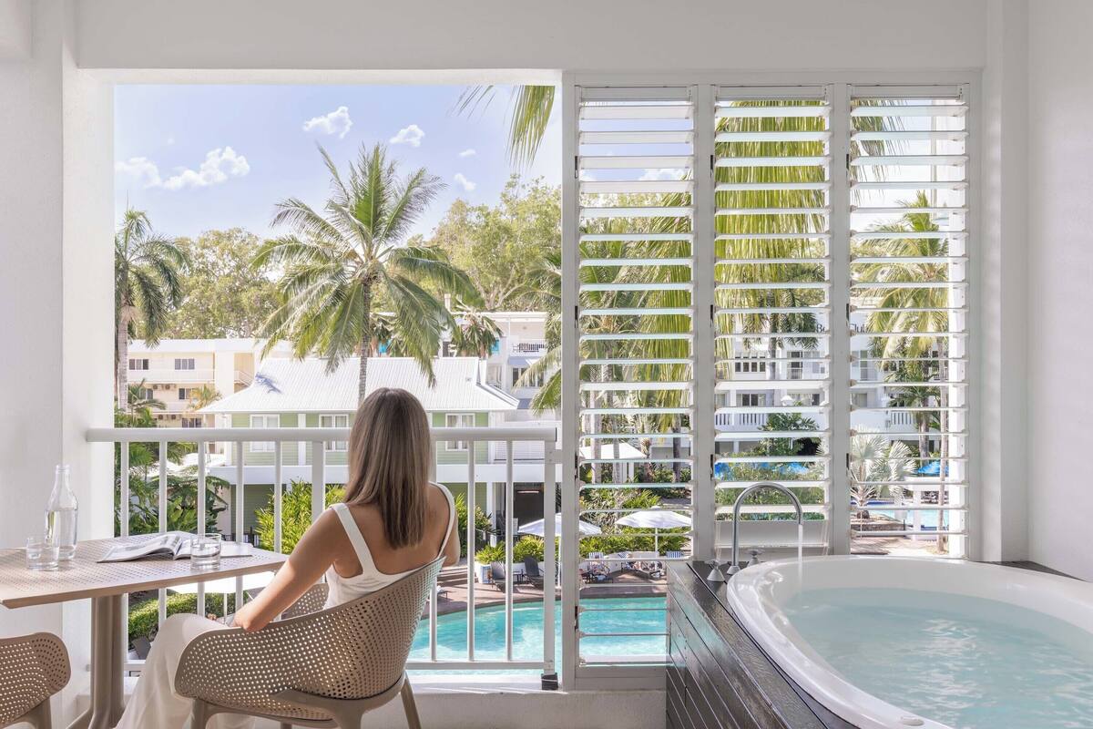 Une femme est assise à une table près d’une fenêtre donnant sur une piscine et un paysage tropical.