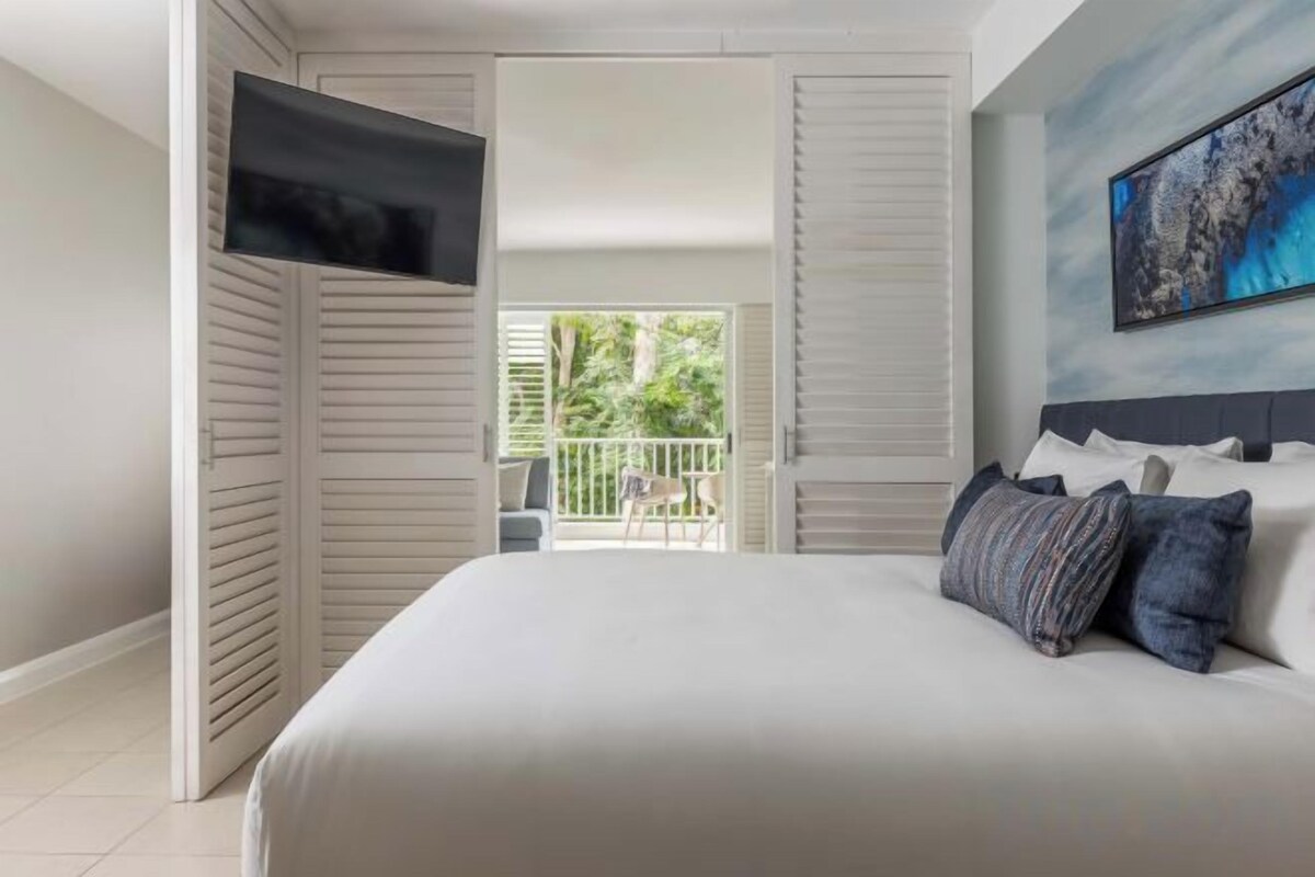 Un dormitorio con cama, televisión montada en la pared y balcón con vistas a la vegetación.