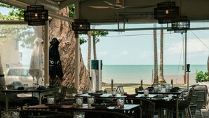 Un restaurante con terraza al aire libre y vistas al mar, con mobiliario de mimbre e iluminación colgante.