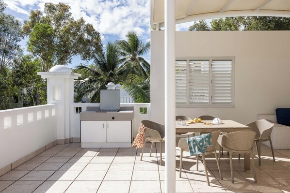 Un patio techado con zona de comedor, paredes blancas y vistas a palmeras.