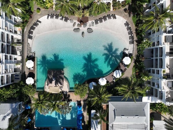 Une vue aérienne d’un complexe hôtelier avec une piscine centrale entourée de chaises longues et de palmiers, et des bâtiments d’hôtel adjacents.