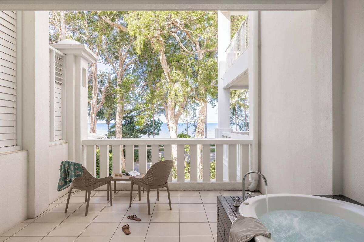 Un balcon avec vue sur les arbres et un plan d’eau, une baignoire et deux fauteuils en osier.