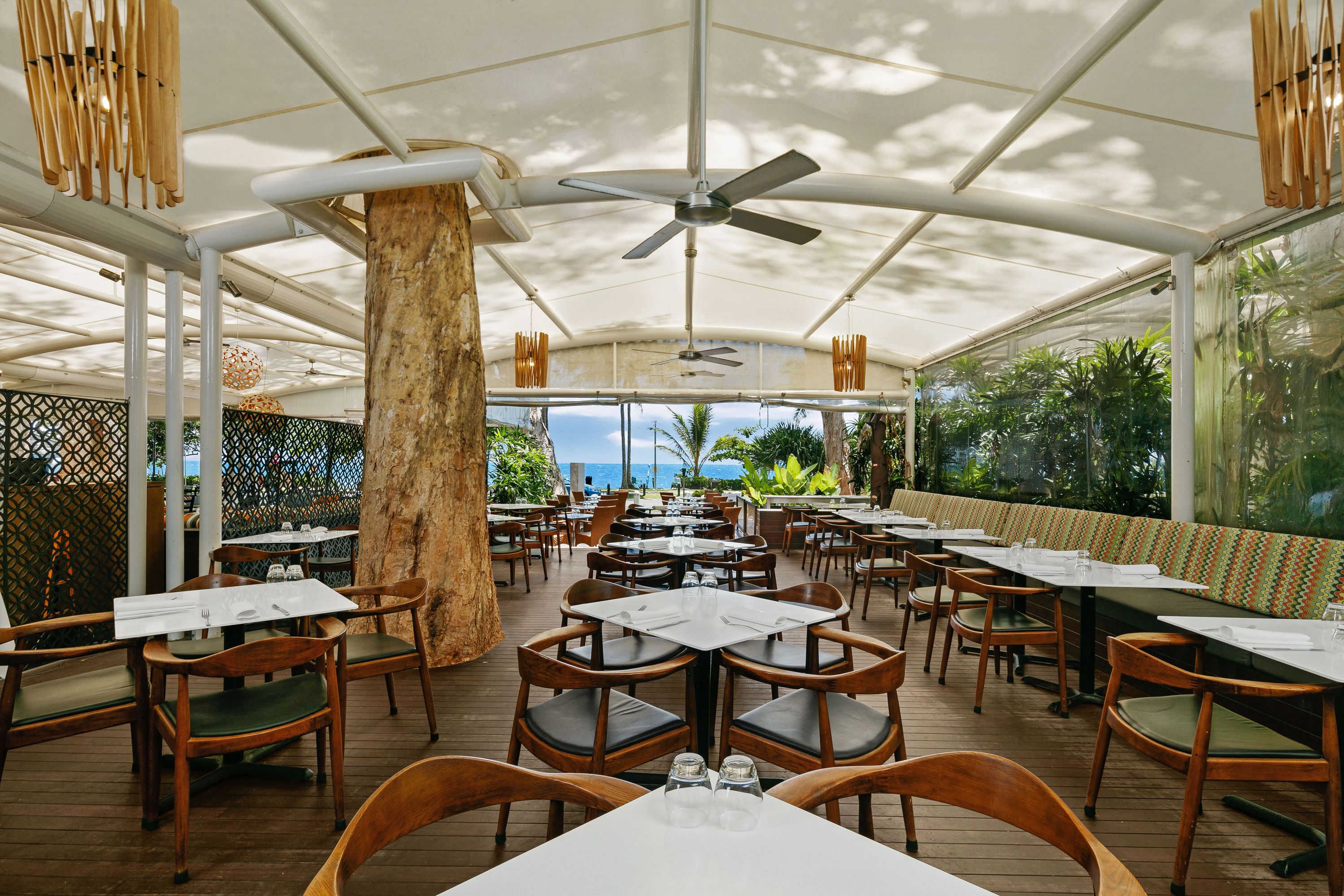 Un restaurant a l'aire lliure amb taules i cadires de fusta, un ventilador de sostre i vista a l'oceà.