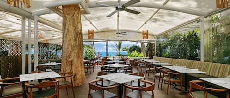An outdoor restaurant with wooden tables and chairs, a ceiling fan, and a view of the ocean.