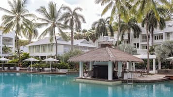 A resort pool with lounge chairs and umbrellas, surrounded by palm trees and a thatched-roof structure.