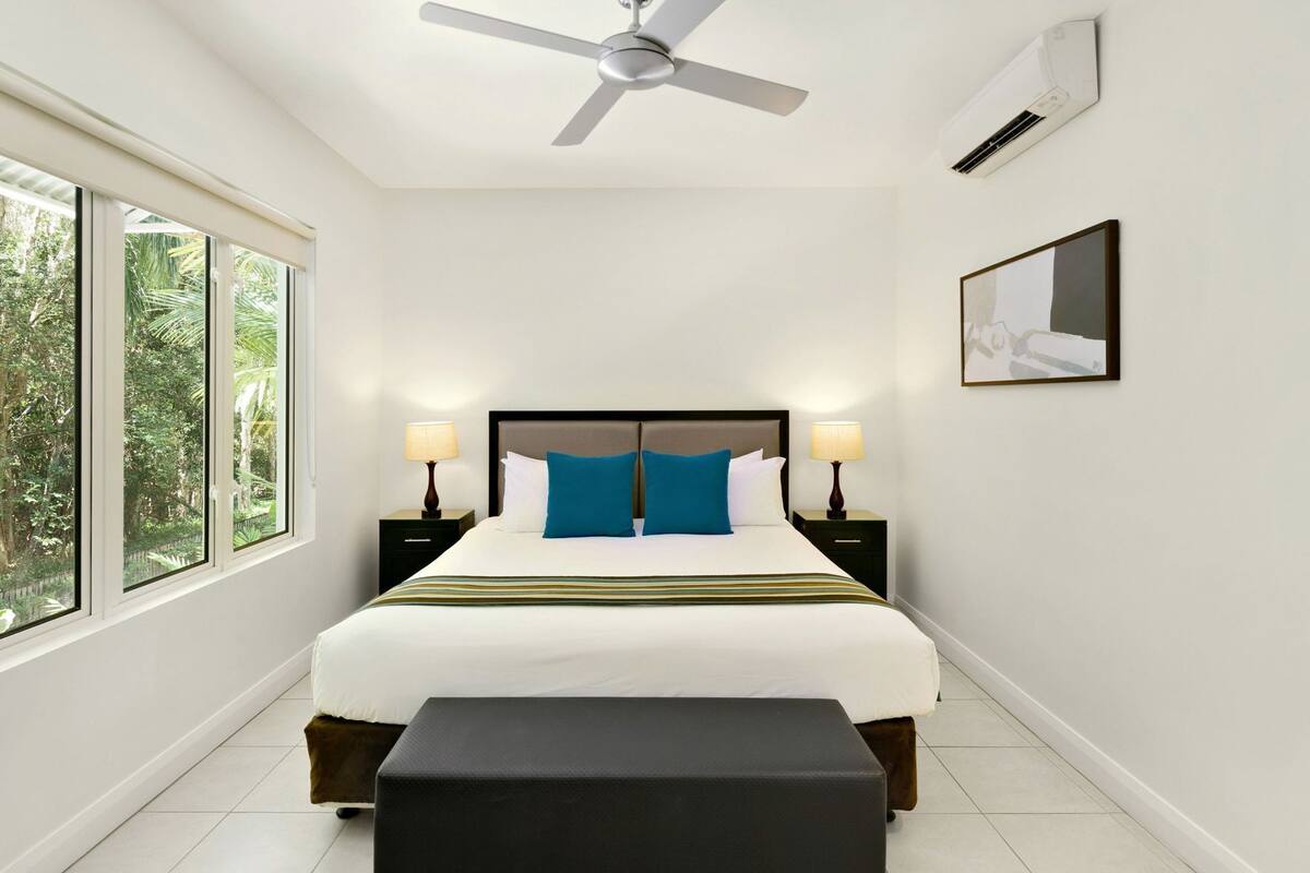 Une chambre d’hôtel équipée d’un lit, d’un ventilateur de plafond, d’une table de chevet avec une lampe, d’une fenêtre donnant sur un espace verdoyant et d’un appareil de climatisation.