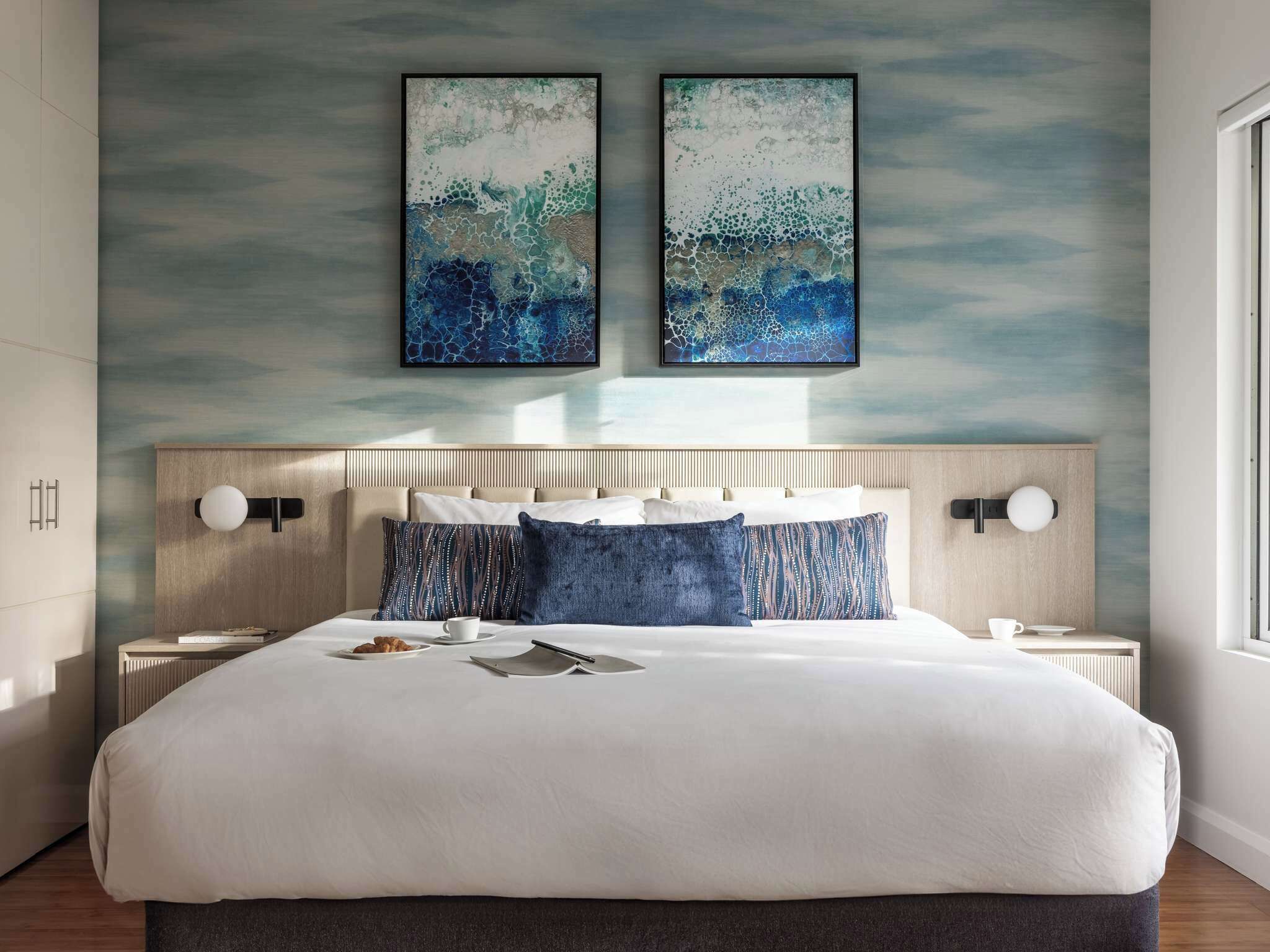 A modern hotel room with a large bed, two bedside tables, and abstract wall art.