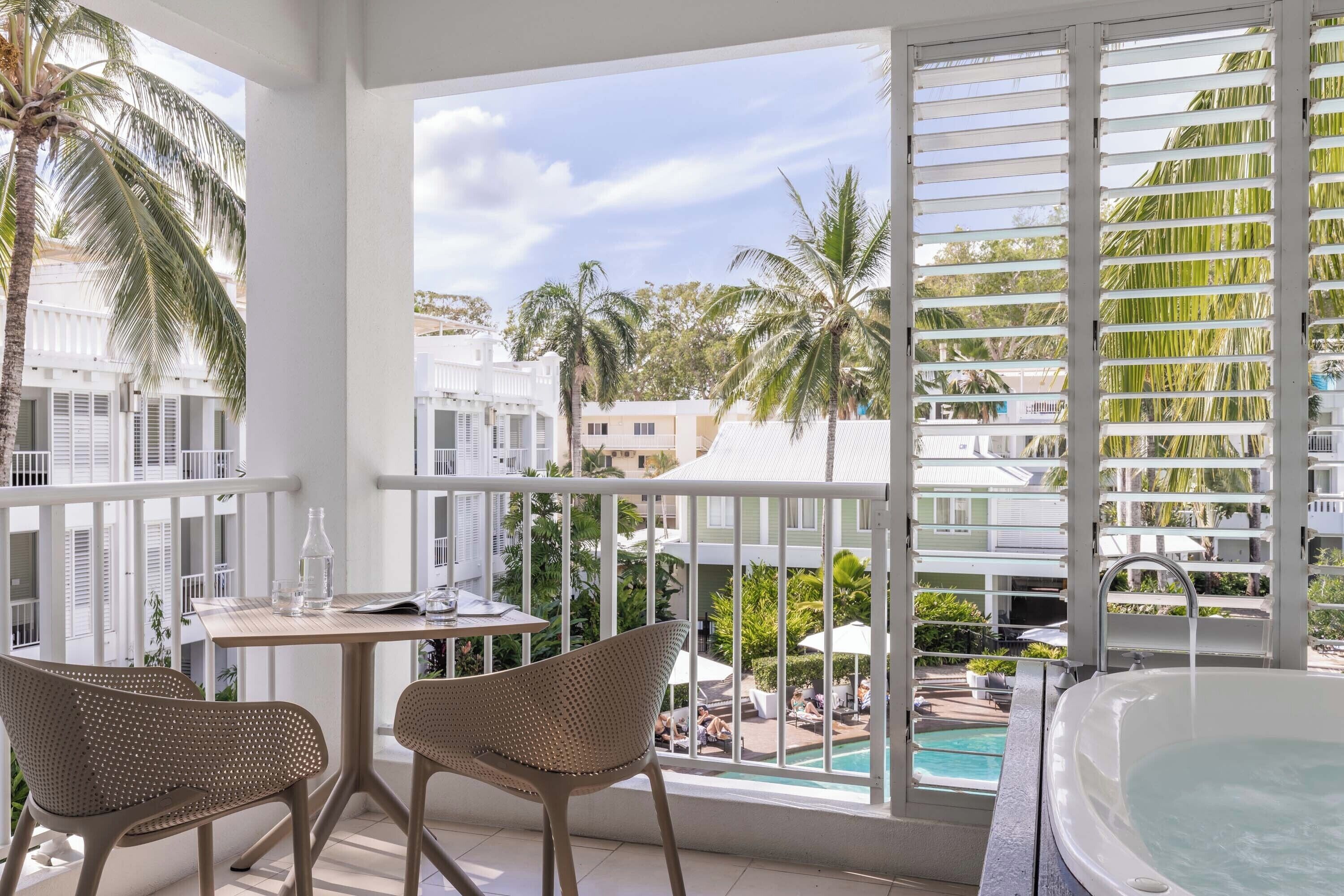 A balcony with a table and two chairs, overlooking a swimming pool and palm trees.