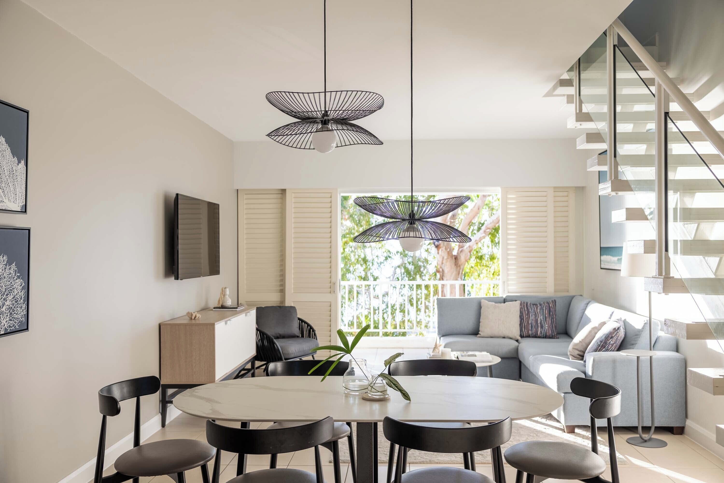 A modern living room with a dining area, featuring a staircase, a sofa set, and pendant lights.