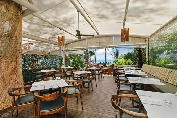 An outdoor restaurant with wooden tables and chairs, a covered seating area, and a view of the ocean.