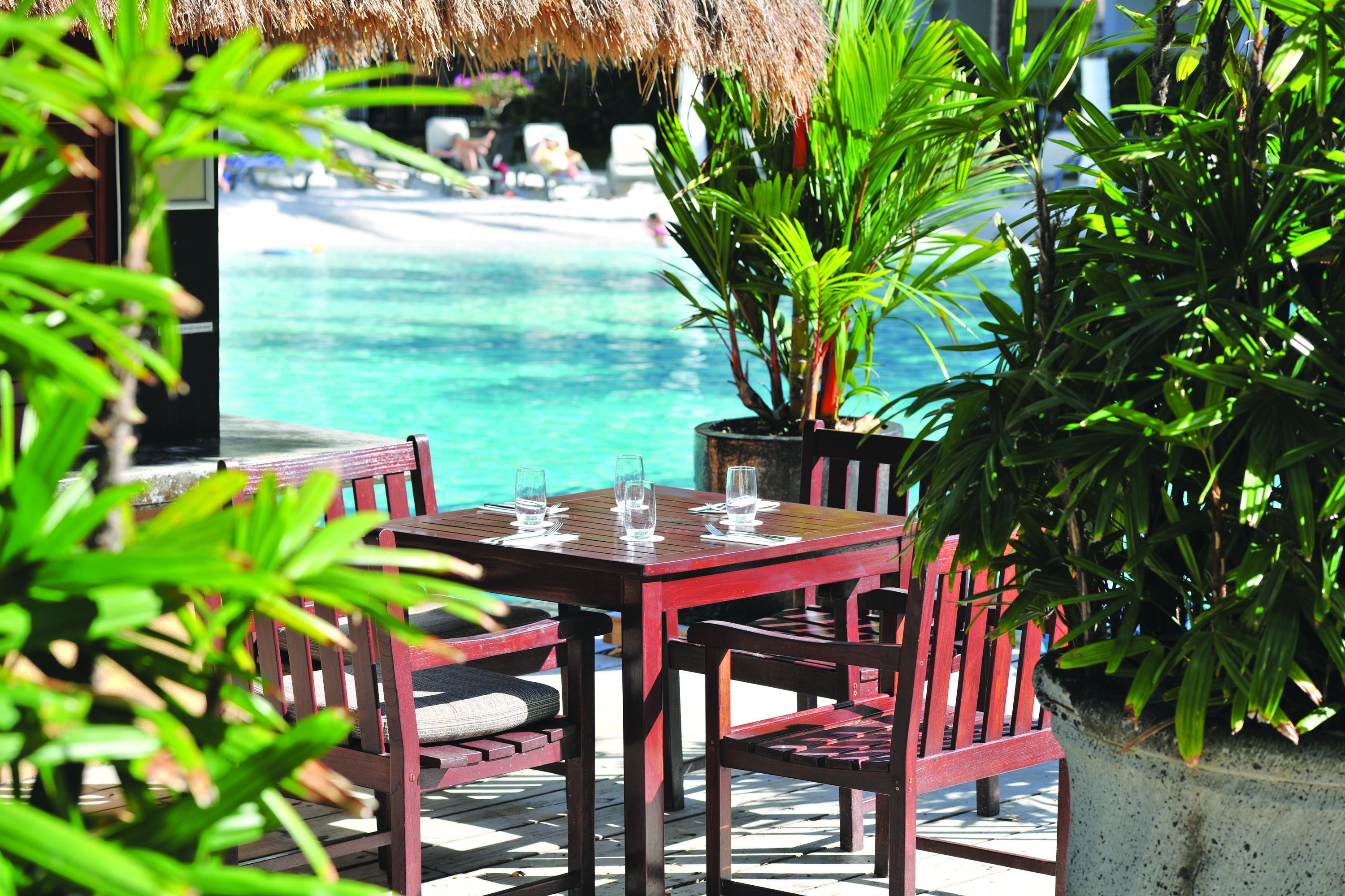 A wooden table with chairs and a glass of water, overlooking a clear blue pool and a beach.