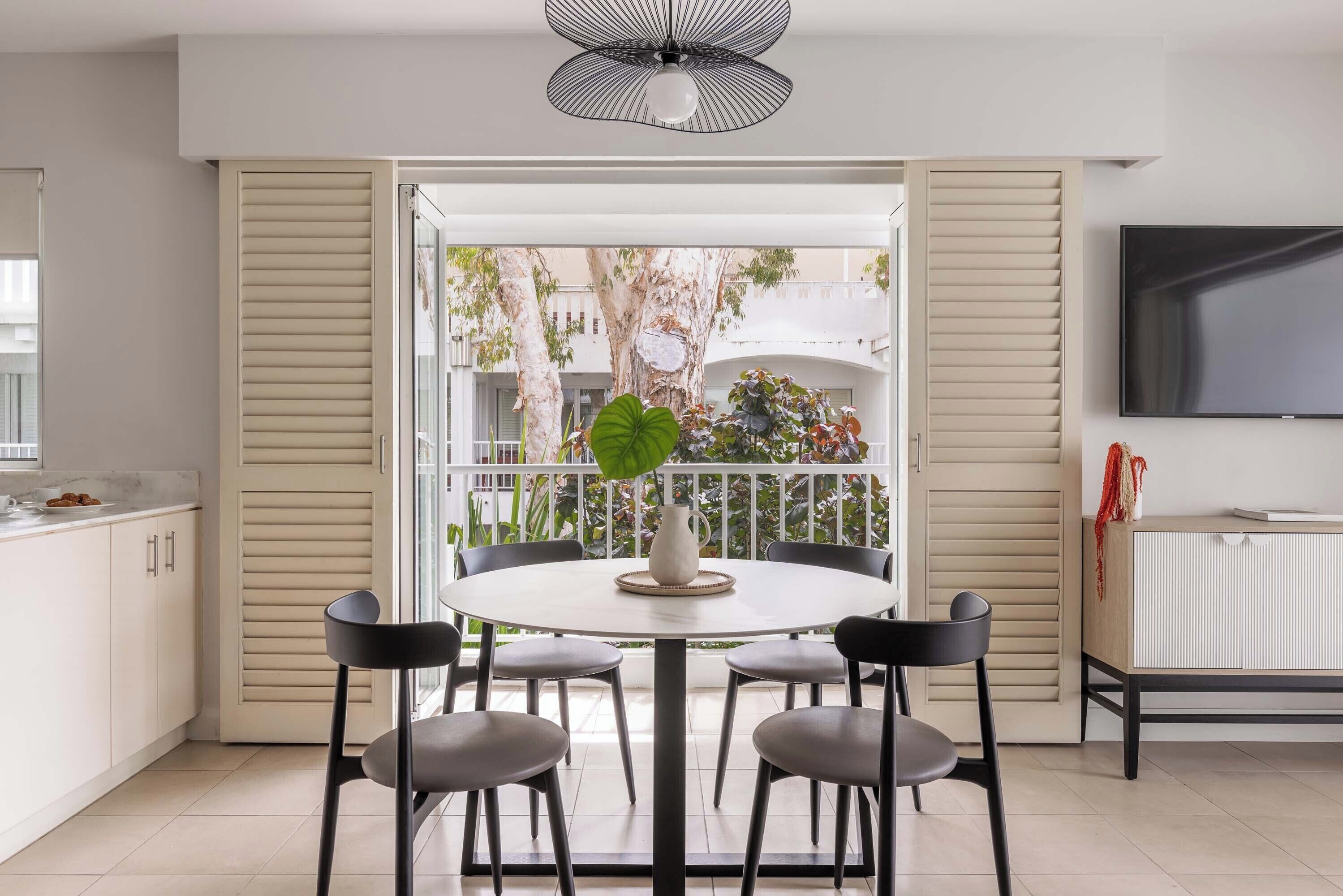A modern kitchen with a dining area, featuring a round table with four chairs, a TV mounted on the wall, and a large window with a view of trees and buildings.