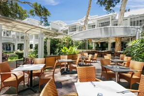 Un espace extérieur aménagé en terrasse, avec des meubles en osier et des tables blanches, entouré de bâtiments blancs et de verdure.