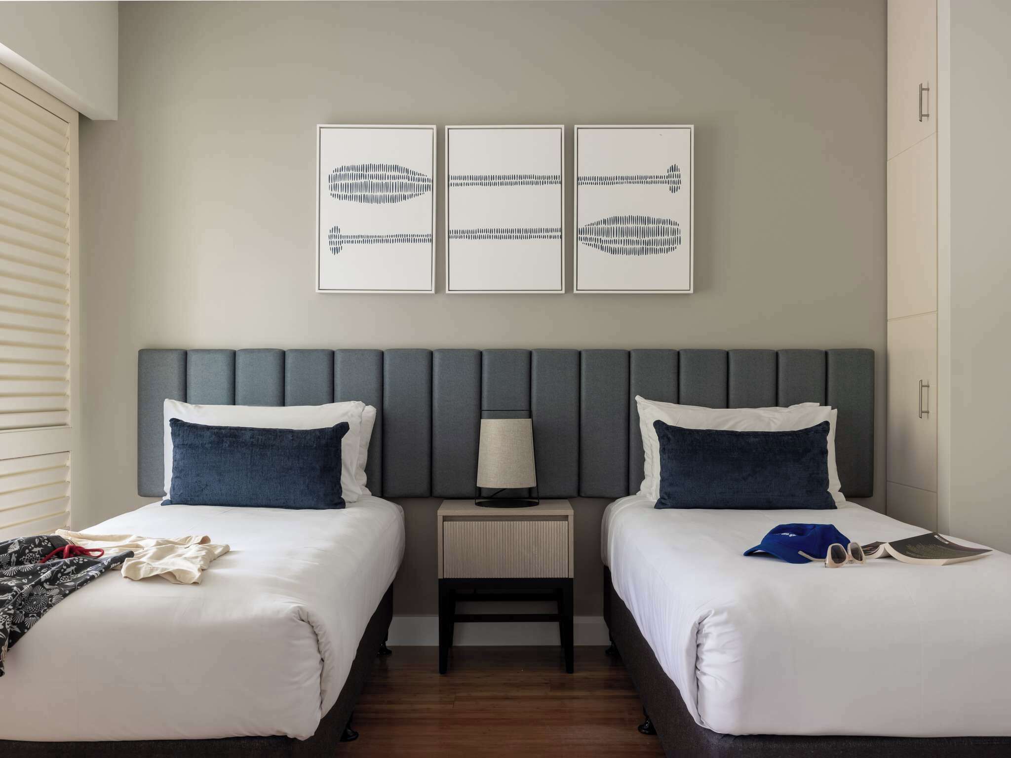 A hotel room with two beds, a grey headboard, a bedside table with a lamp, and three framed artworks on the wall.
