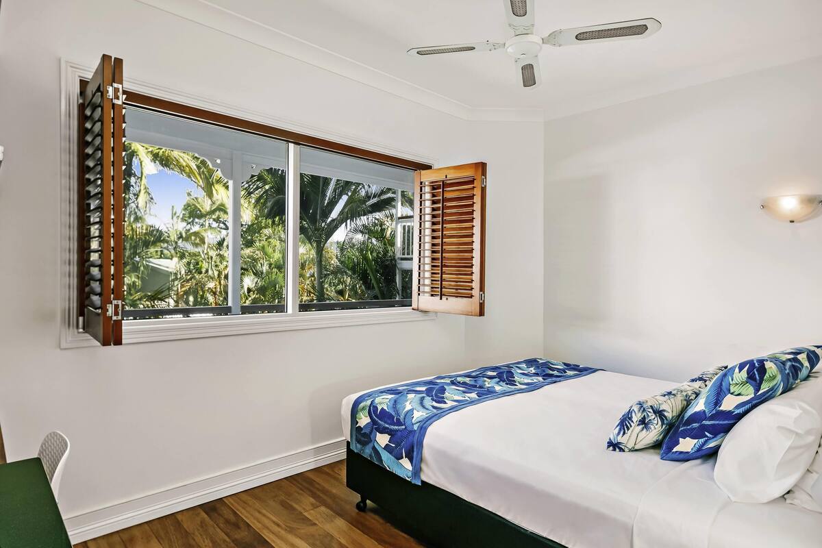 A bedroom with a bed, a window with shutters, a ceiling fan, and a view of palm trees.