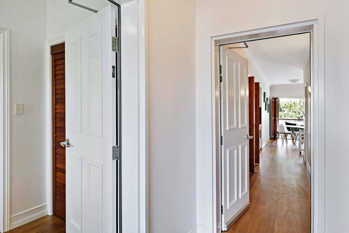A hallway with white walls and wooden flooring, leading to a dining area with a table and chairs.