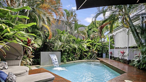 A pool with a waterfall, surrounded by lush tropical plants and wooden deck seating.