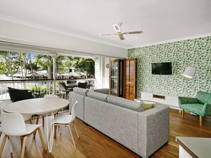 A modern living room with a large grey sofa, a dining table with chairs, a TV mounted on the wall, and a balcony with outdoor furniture. - Mantra On The Inlet (Port Douglas)