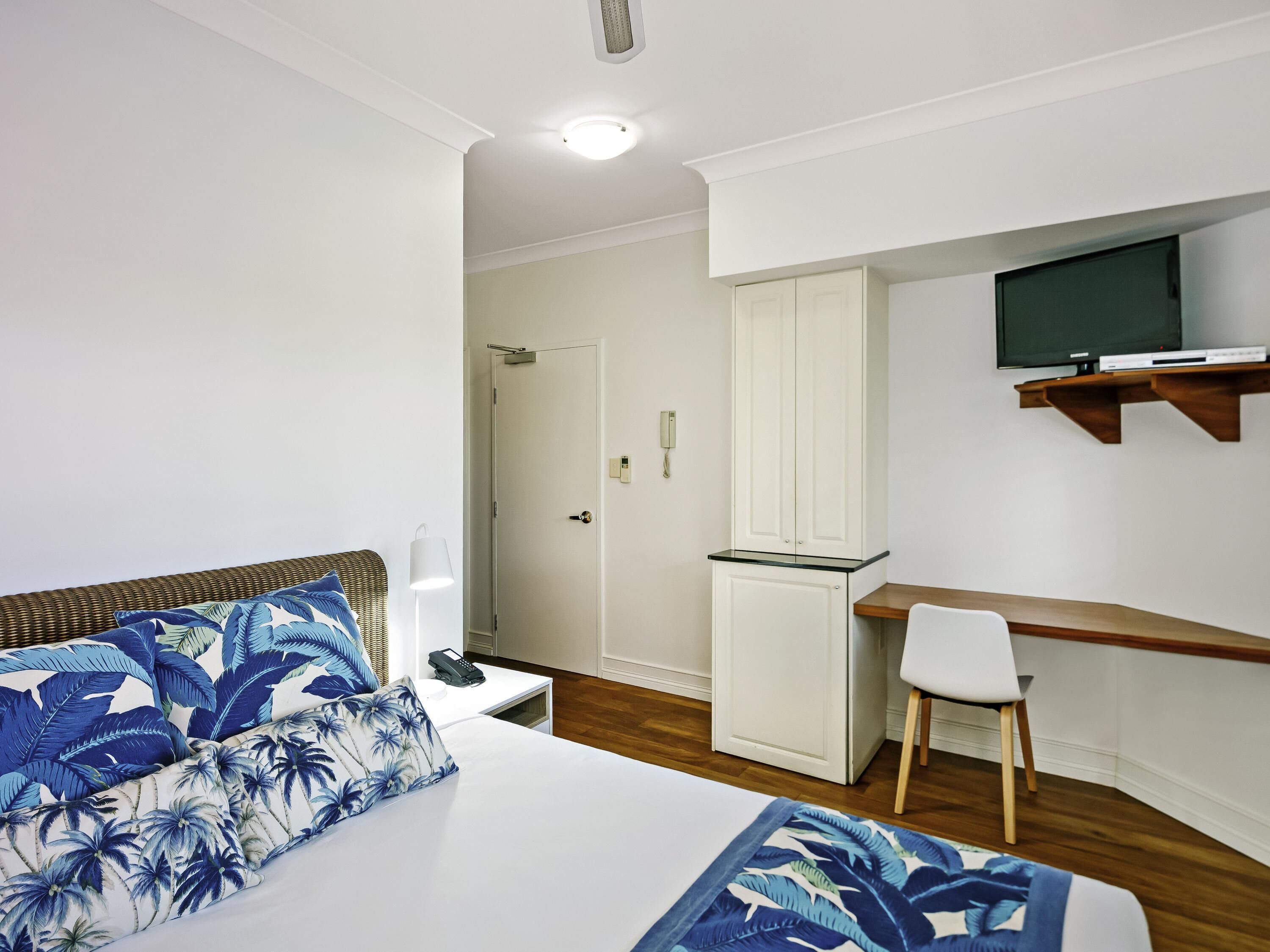 A bedroom with a bed, a desk, a chair, a wardrobe, and a television on a shelf.