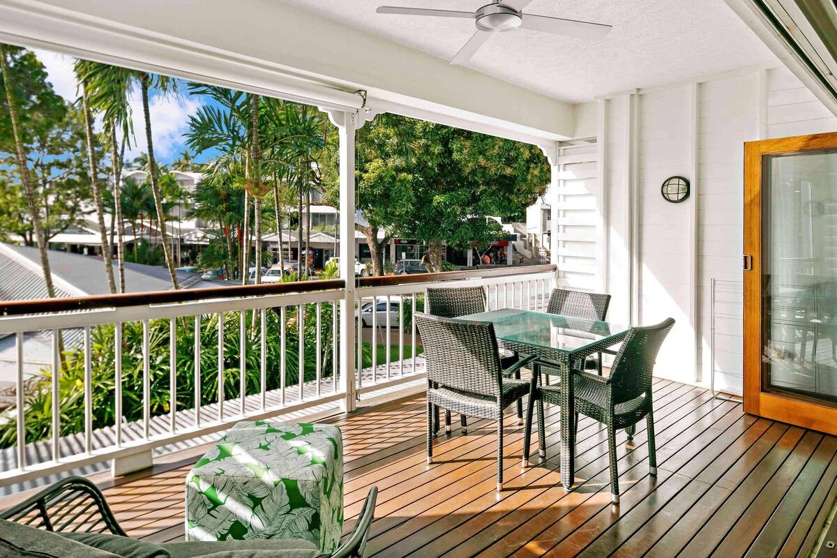 A covered outdoor area with a ceiling fan, a table with chairs, and a view of palm trees and buildings.