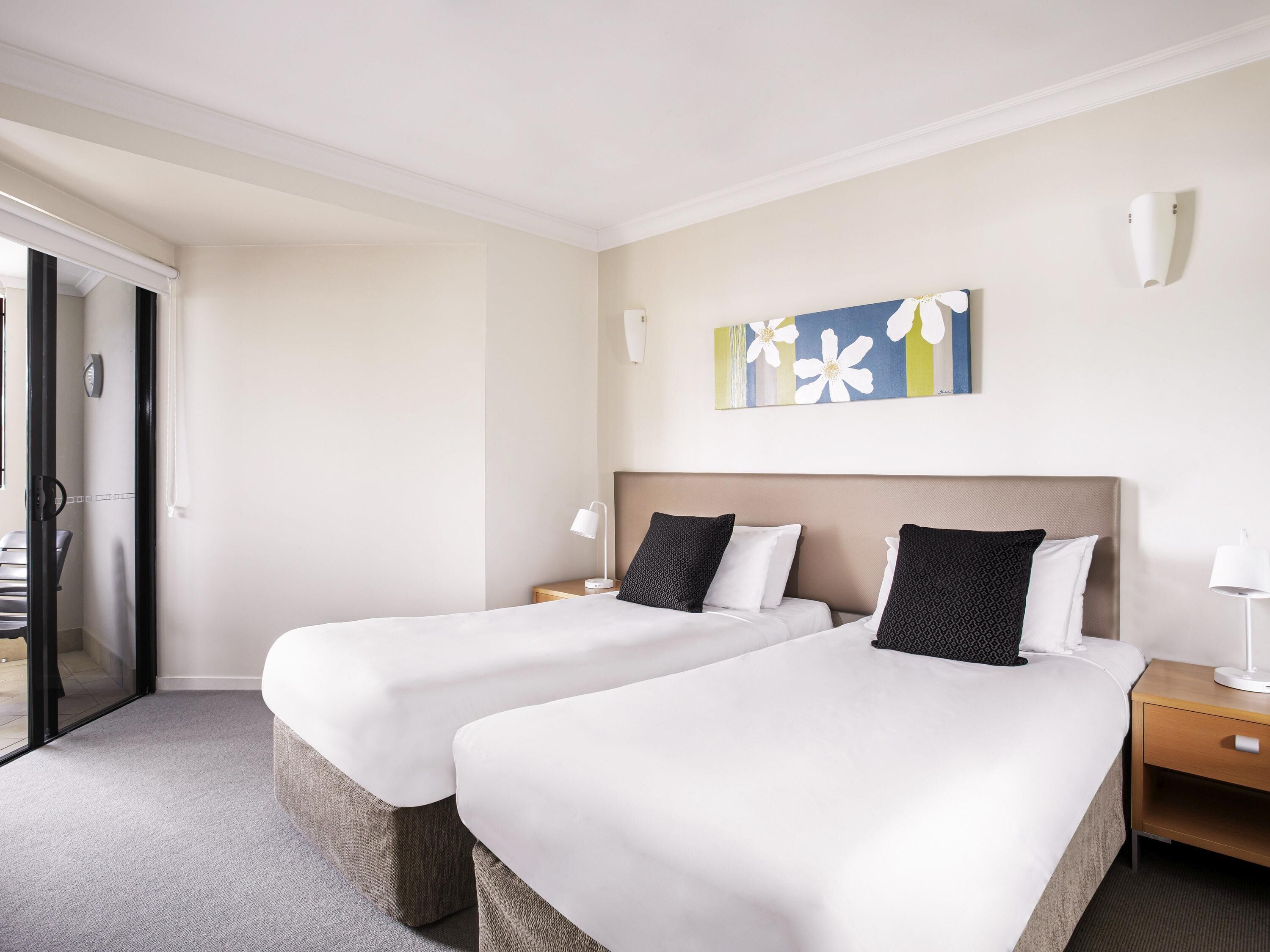 A hotel room with two beds, a small table, a nightstand, a painting, and a door leading to a balcony.