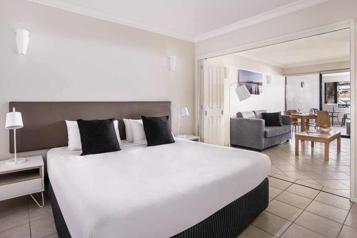 A hotel room with a large bed, a sofa, a dining table, and a view of the outside.