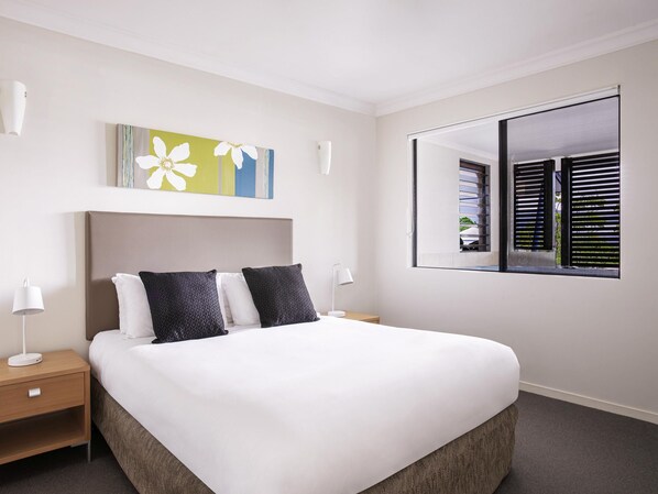 A hotel room with a large bed, two bedside tables, a nightstand, a window with blinds, and a wall-mounted artwork. - Mantra Heritage (Port Douglas)